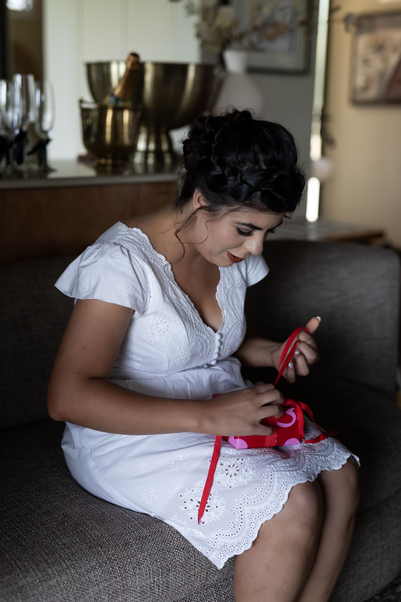 A person in a white eyelet dress sits on a grey couch examining a small red item.