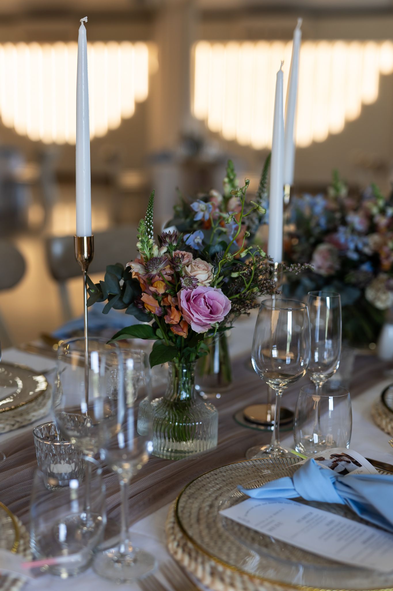 Elegant wedding table setting with tall white candles and pink rose centerpiece in vintage glass vase.