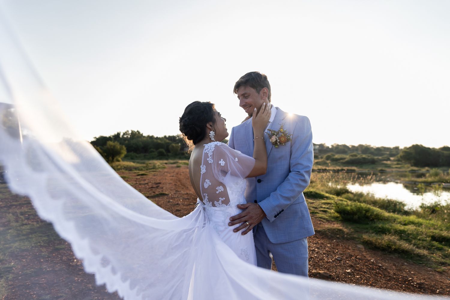 A couple poses dramatically with flowing white dress fabric against a lakeside landscape.