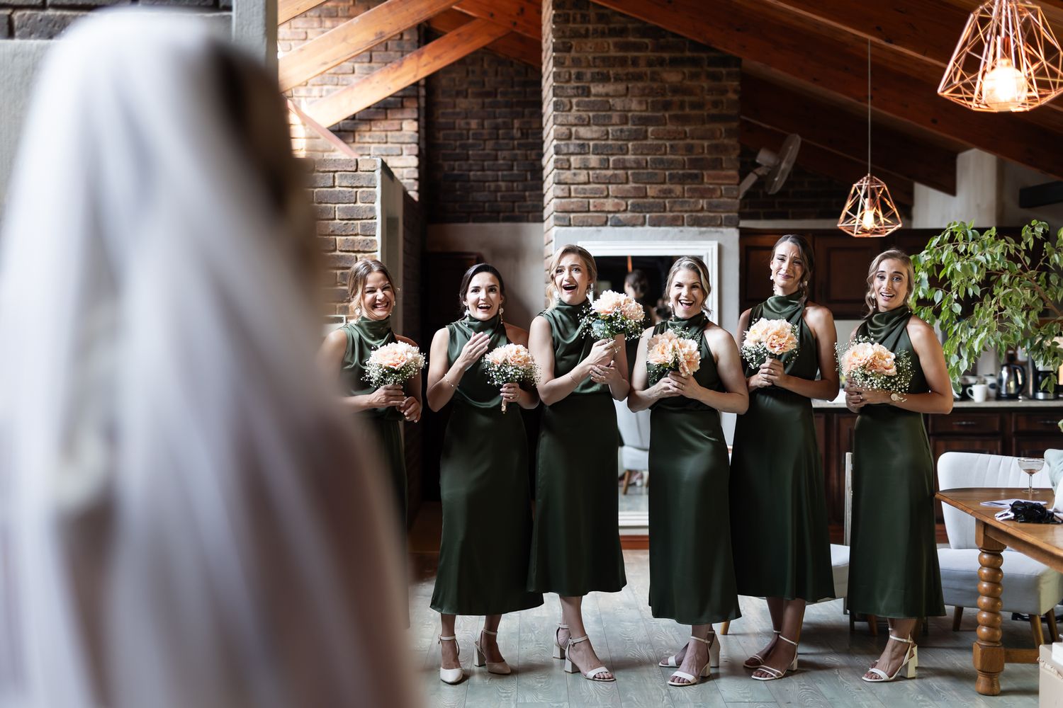 Bridesmaids in elegant emerald green dresses stand together holding bouquets in a rustic brick-walled venue.