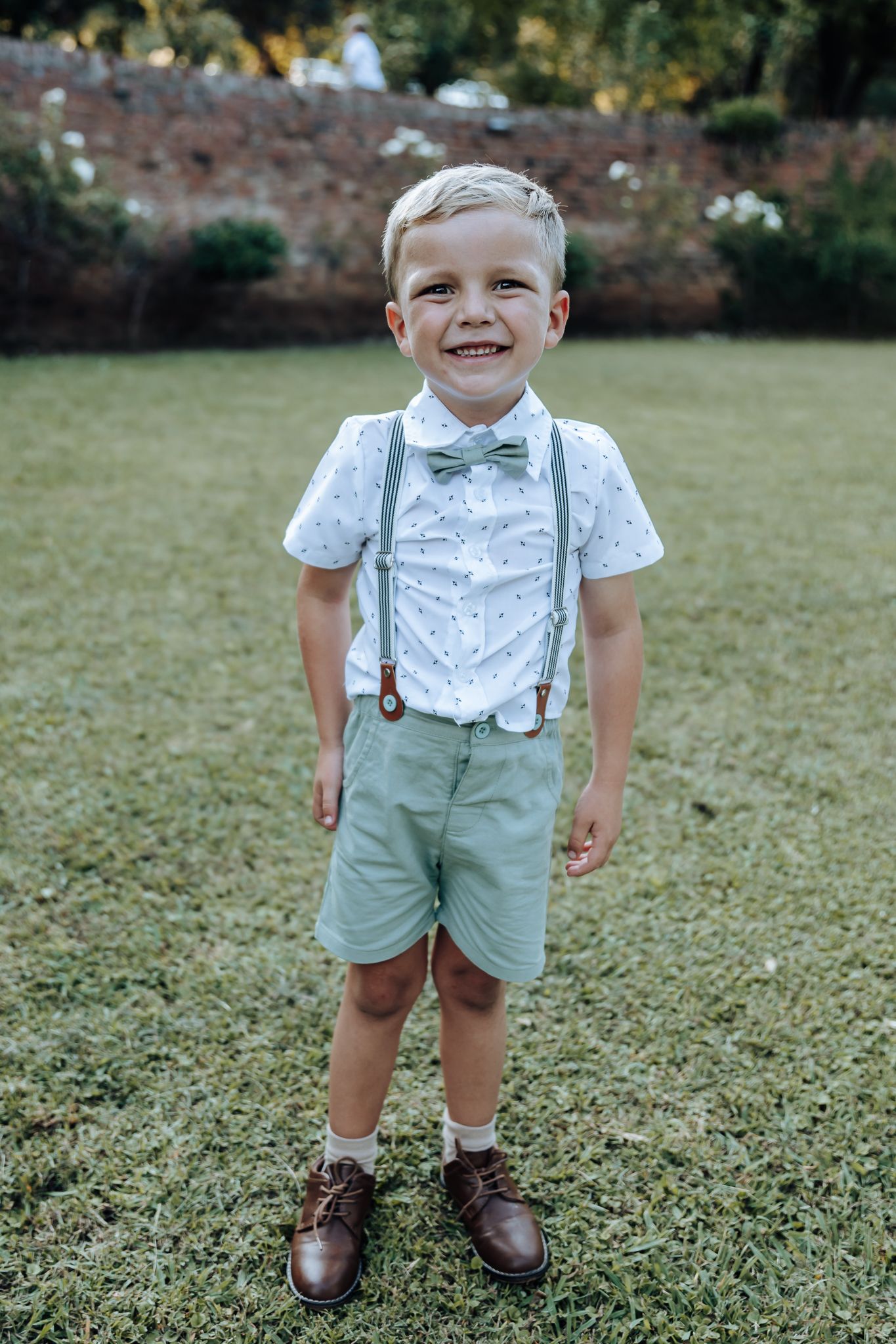 Adorable boy in formal attire with bow tie and suspenders stands on lawn.