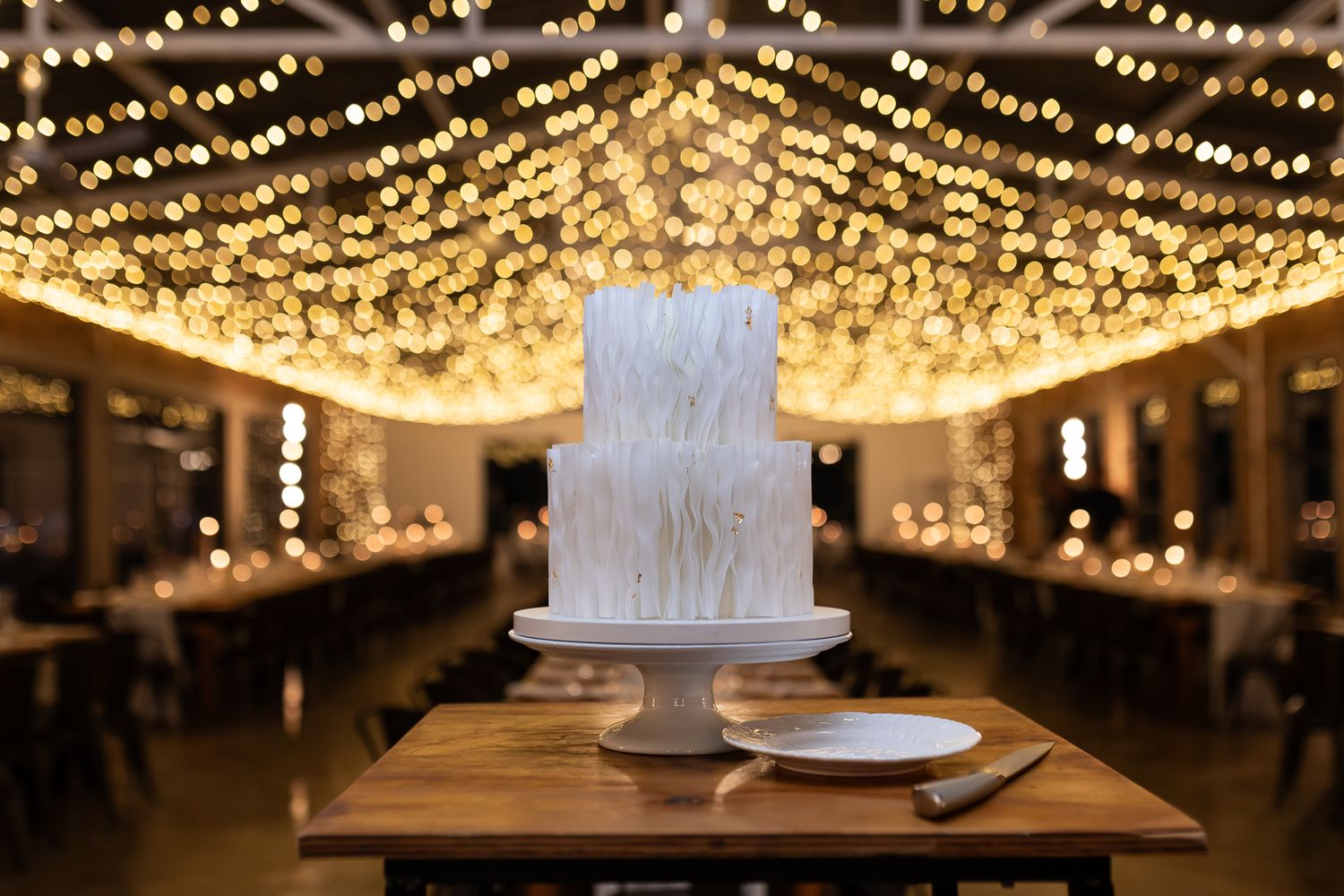 White wedding cake displayed under romantic twinkle light ceiling in rustic reception venue.