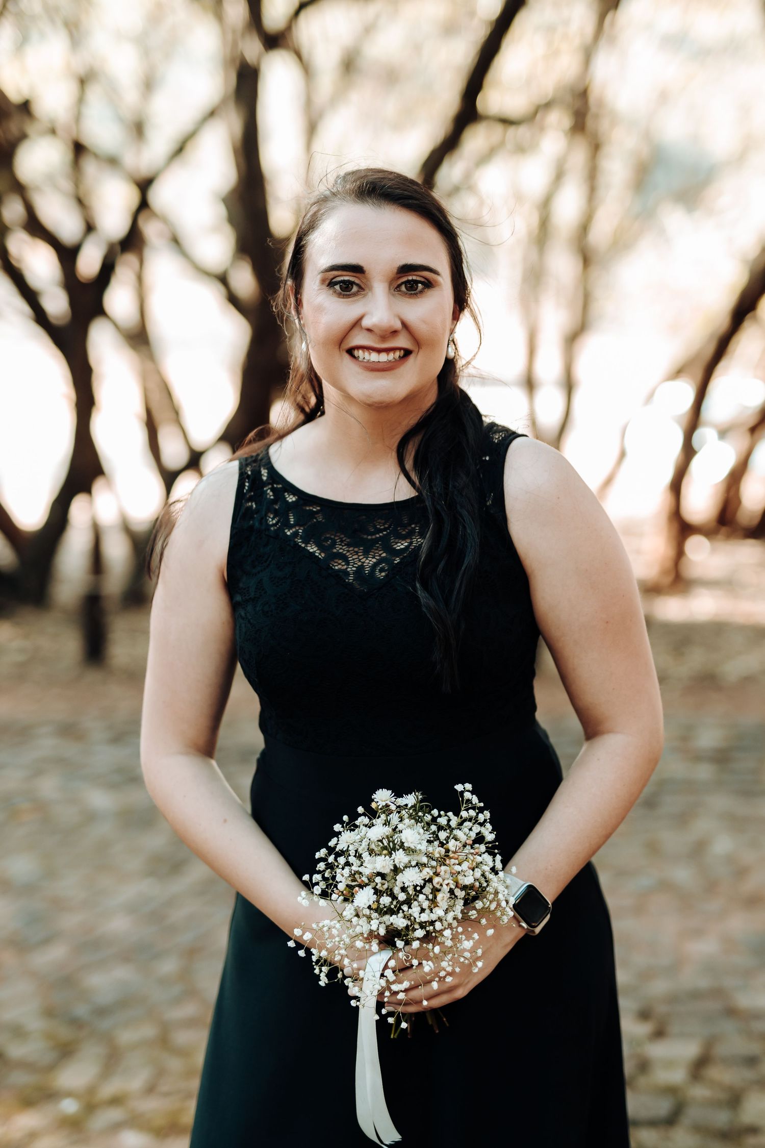 Bridesmaid in sleeveless black lace dress holds white bouquet while smiling outdoors.