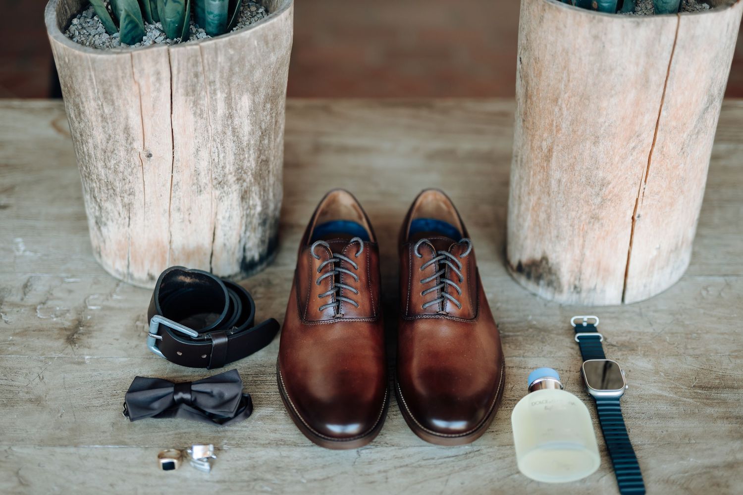 Brown leather dress shoes with accessories including watch and sunglasses on wooden floor.