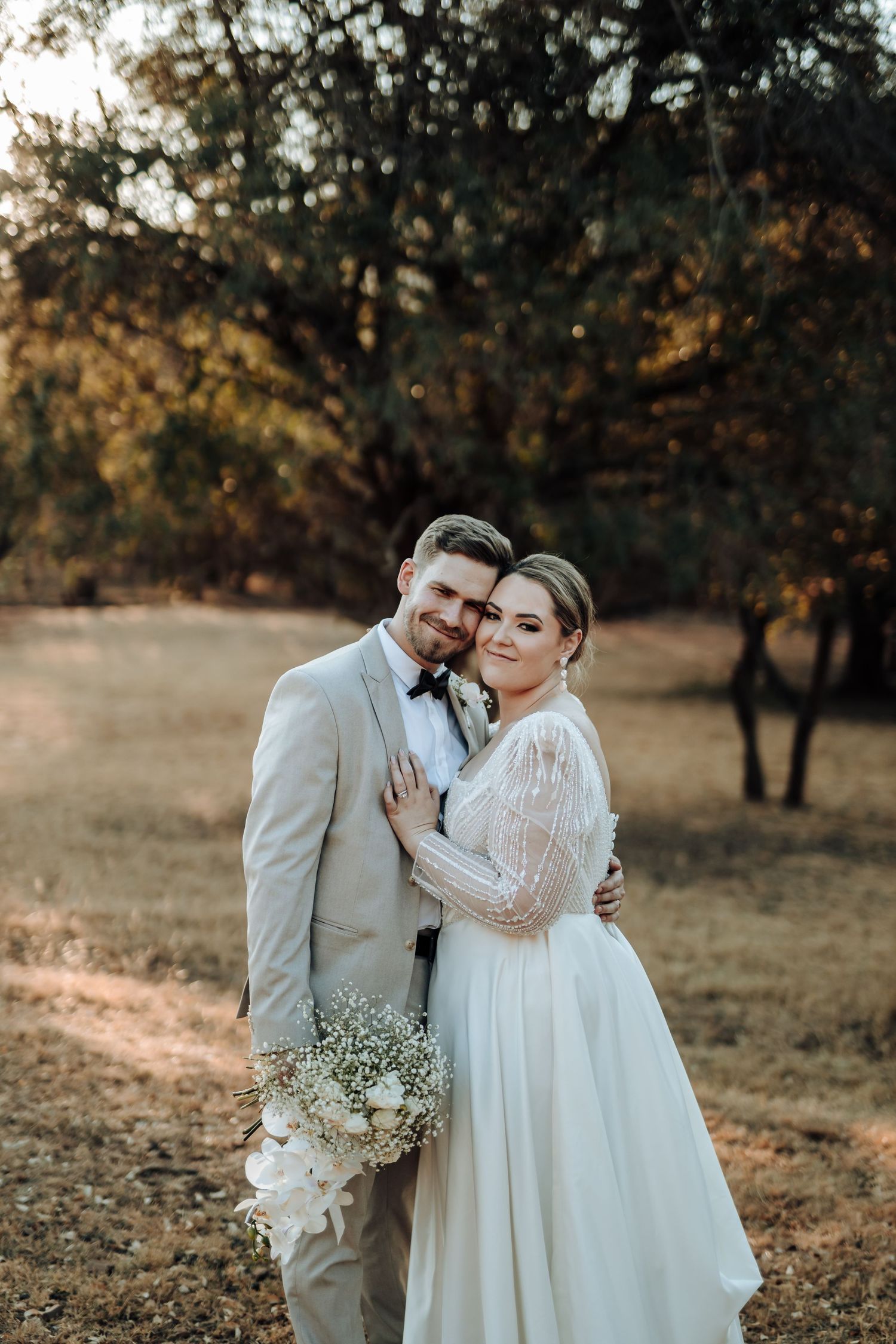 Golden hour wedding portrait with couple holding white bouquet in rustic outdoor setting.