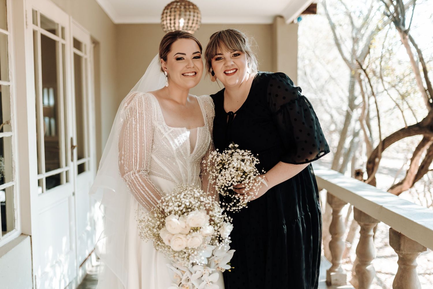 Bride and bridesmaid pose with matching white bouquets under hanging light fixture.