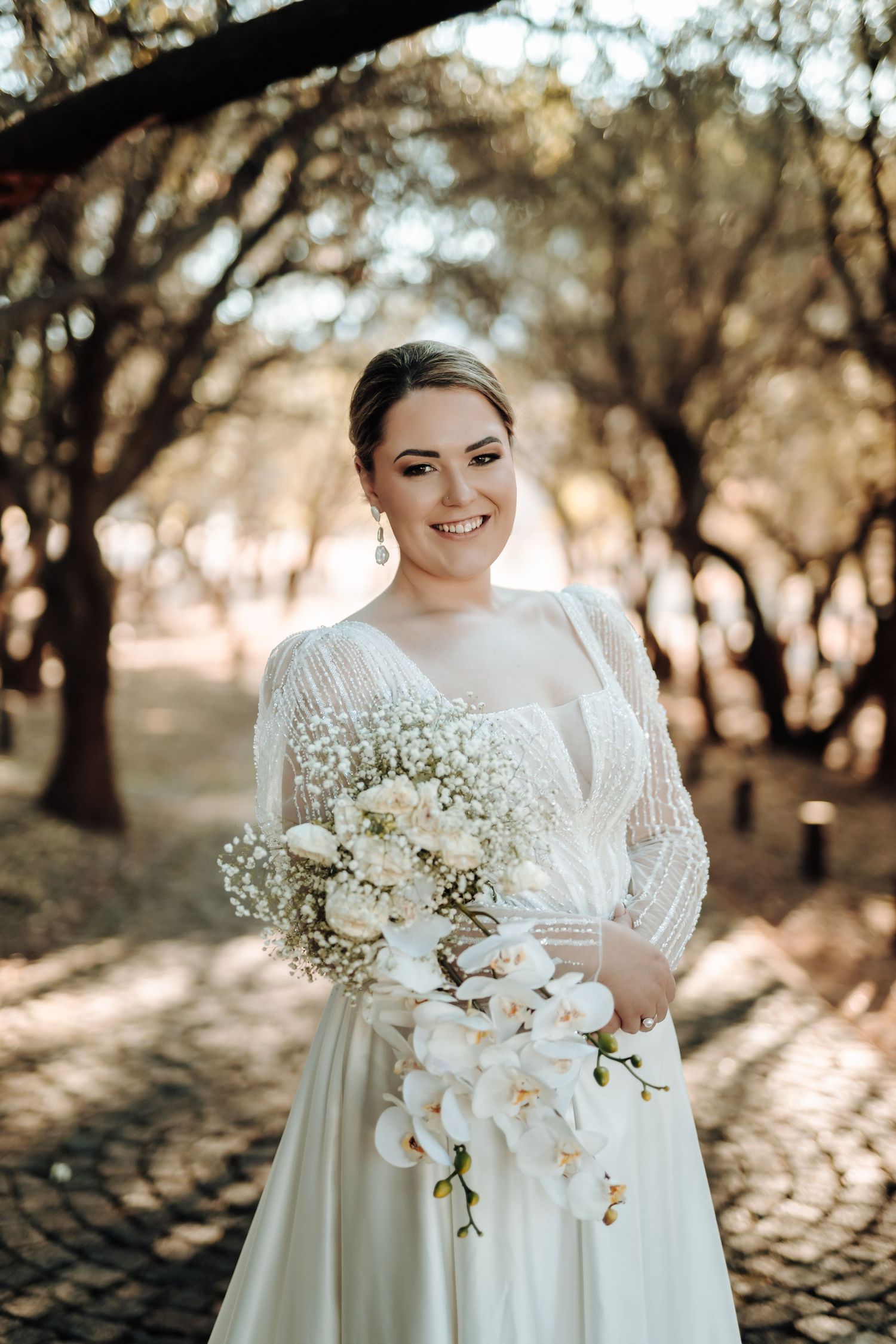 Joyful bride in long-sleeved white gown holds orchid and baby's breath bouquet in wooded setting.