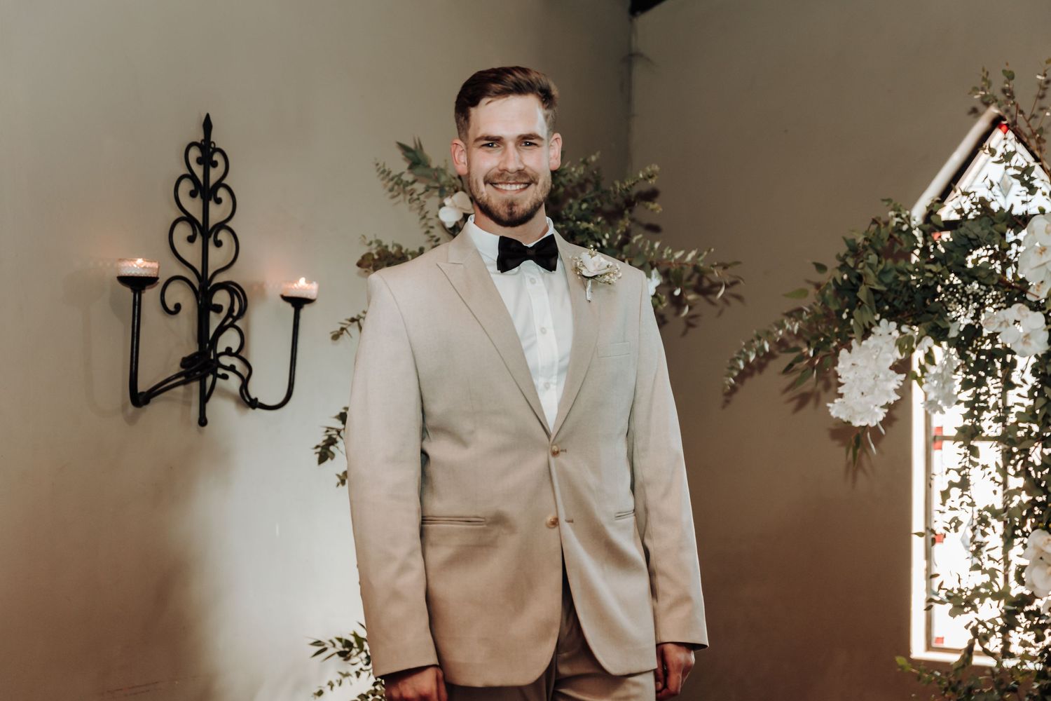 Groom in light beige tuxedo and bowtie stands in chapel setting.