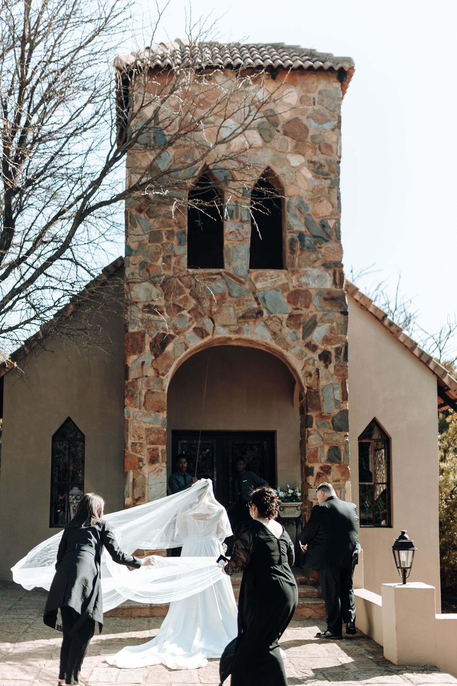 Wedding party helping with bridal veil outside stone chapel with arched windows.