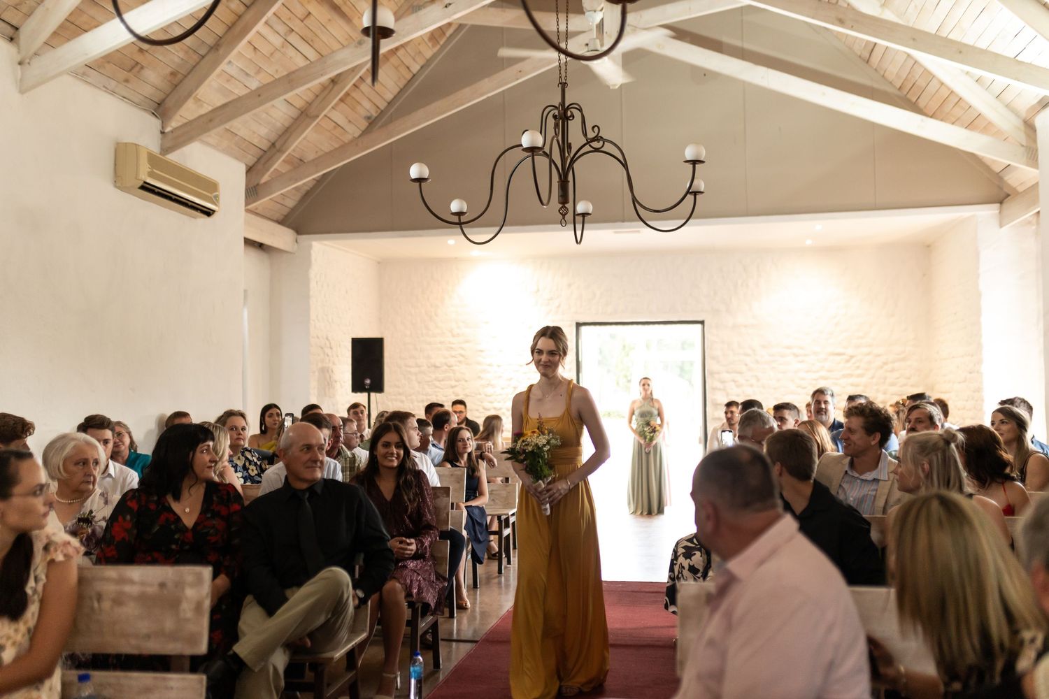Bridesmaid walks down red-carpeted aisle in sage green dress during chapel ceremony.