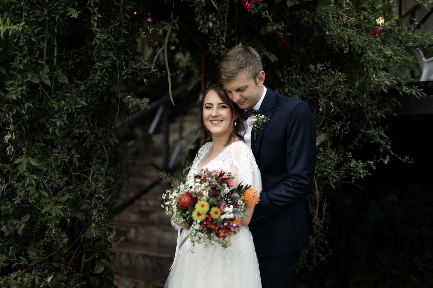 Newlyweds pose with colorful autumn bouquet under flowering vines.
