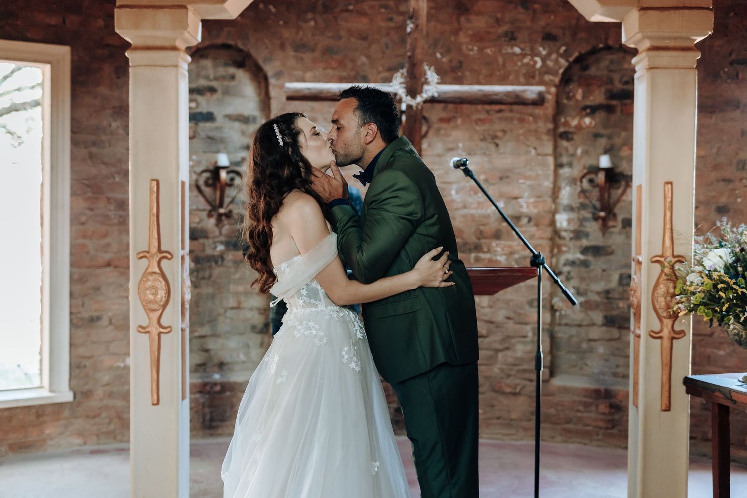 Couple sharing their first kiss as newlyweds under an ornate archway in a brick chapel.