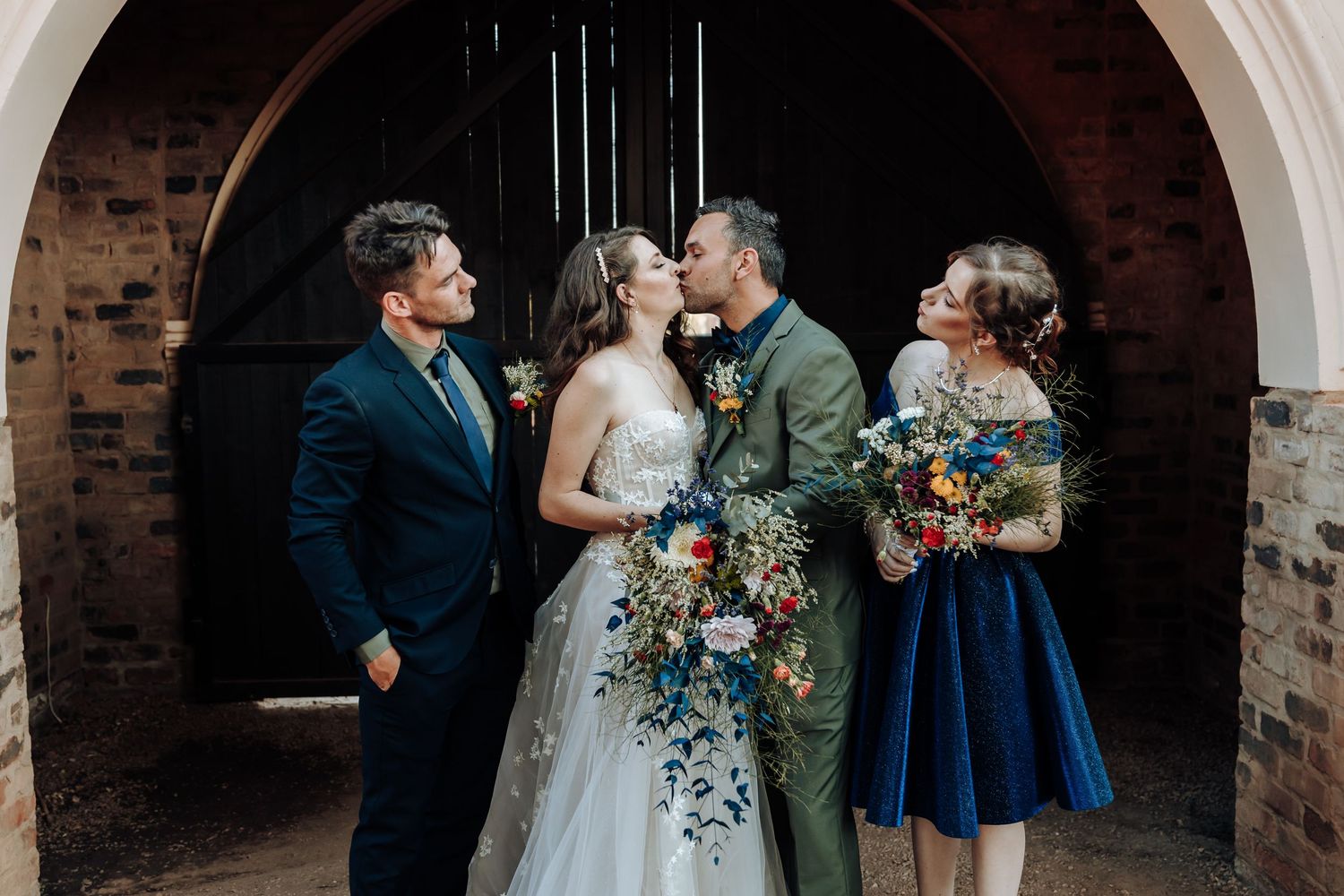 Wedding party shares a candid moment under stone archway with beautiful floral bouquets.