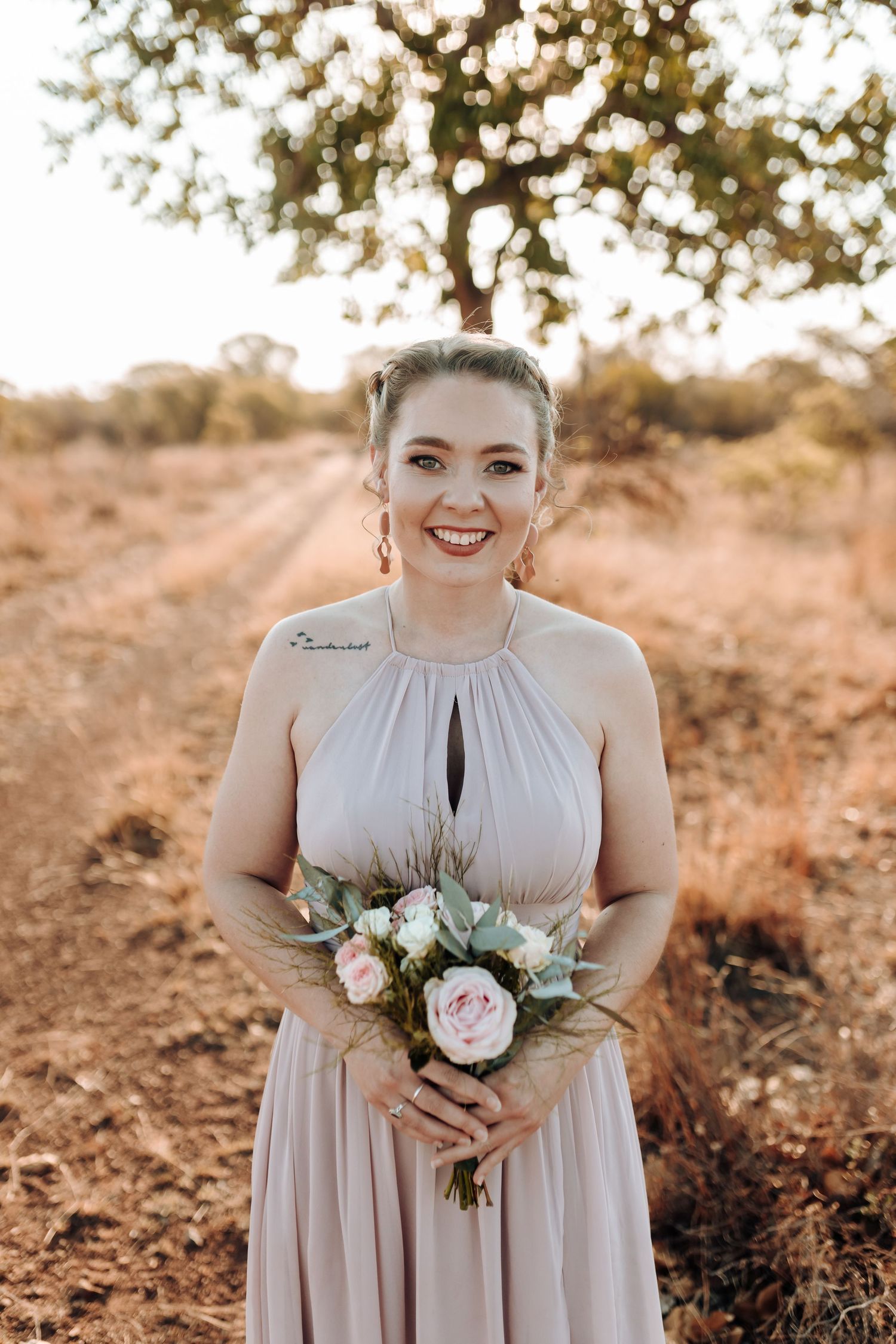 Person in light mauve dress smiling while holding delicate floral bouquet outdoors.