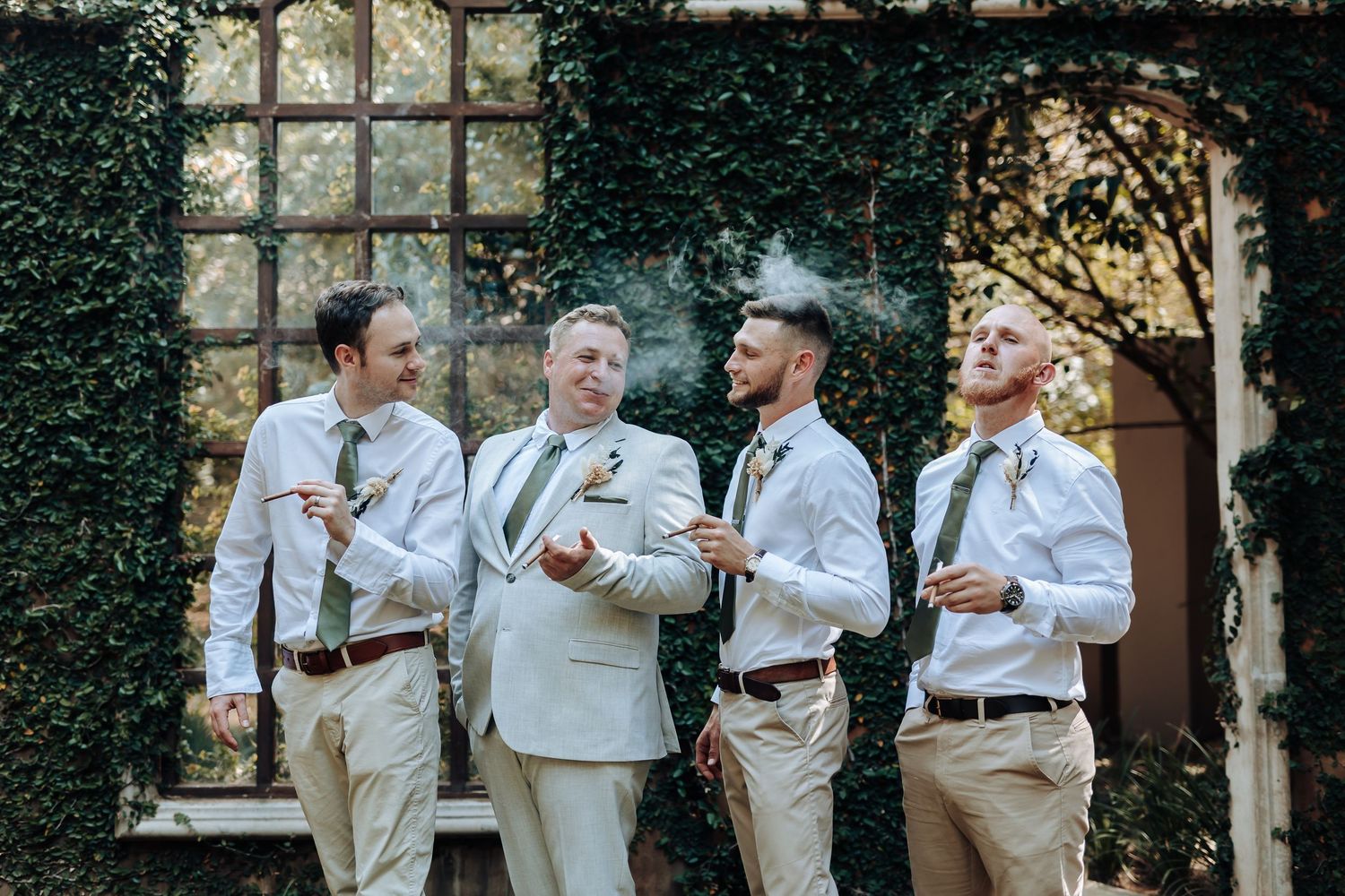 Wedding group enjoys candid moment in coordinated light outfits by ivy-covered wall.