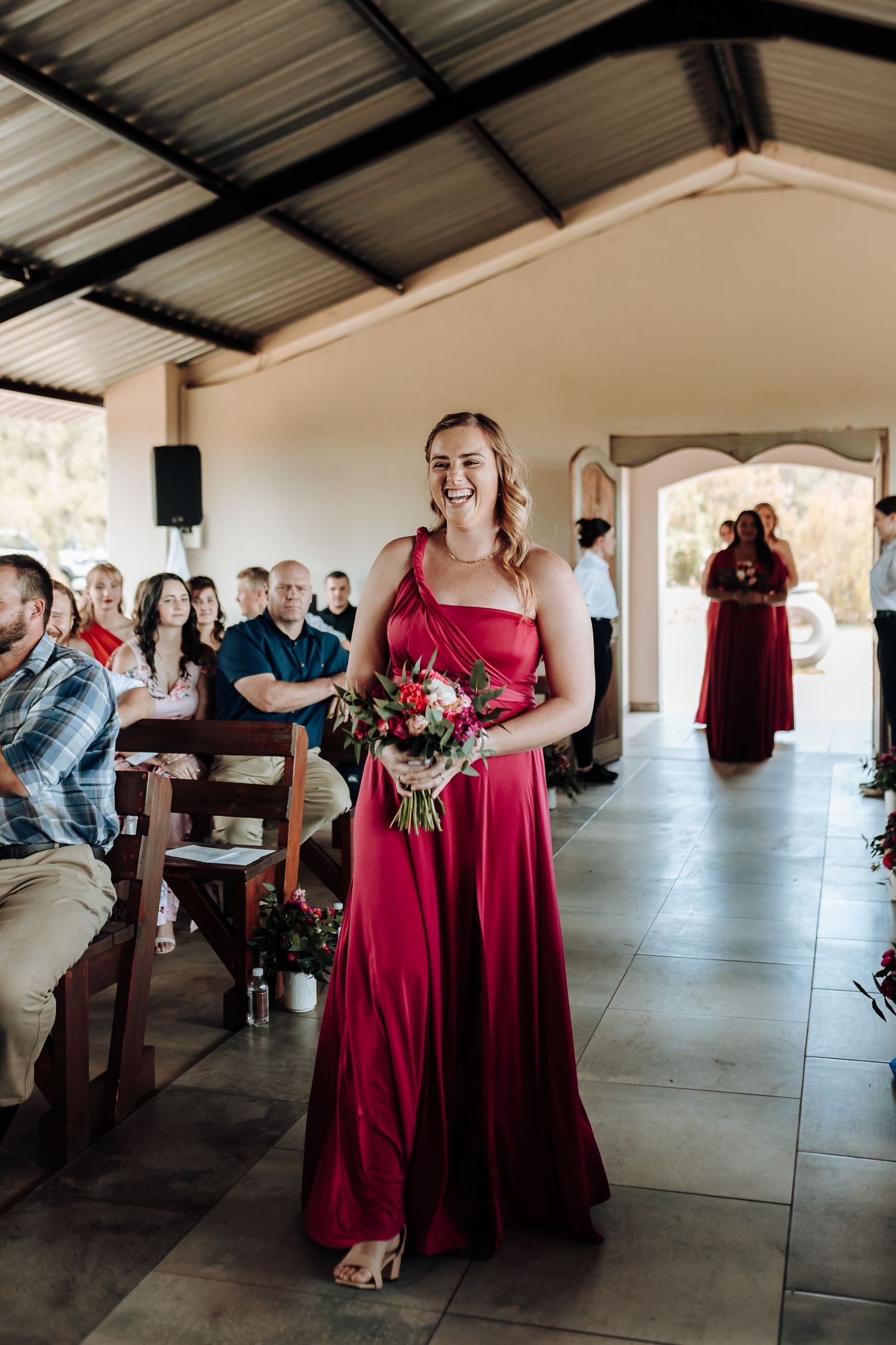 Bridesmaid in flowing red dress holds bouquet while standing in covered outdoor venue.