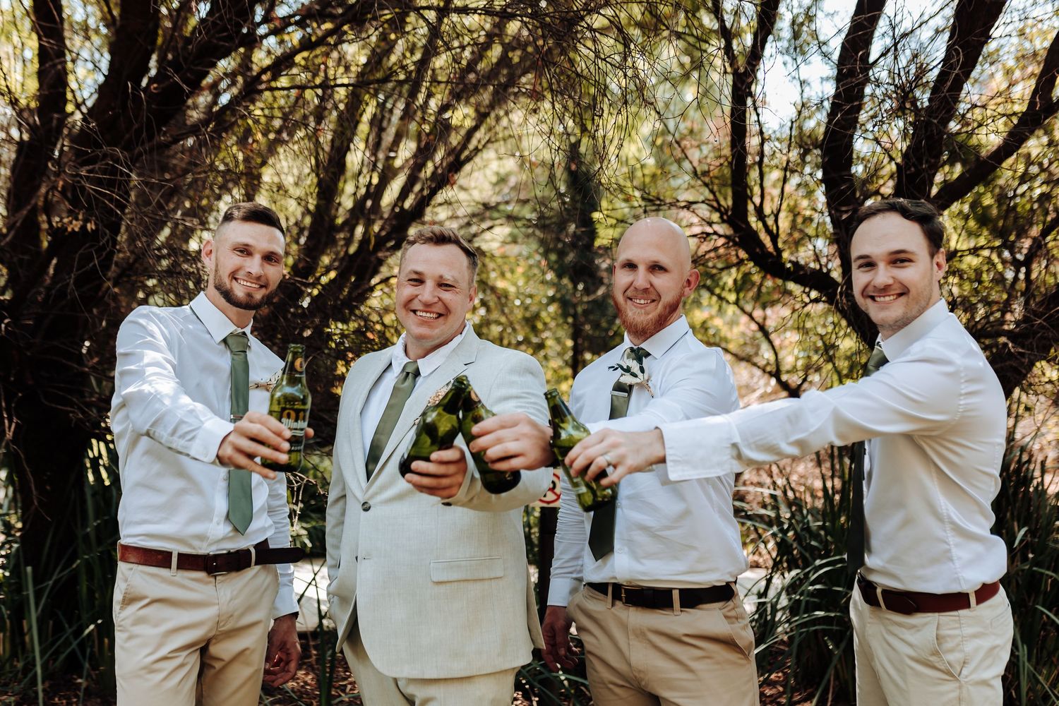 Groomsmen in light colored attire raise glasses for a celebratory toast under tree canopy.