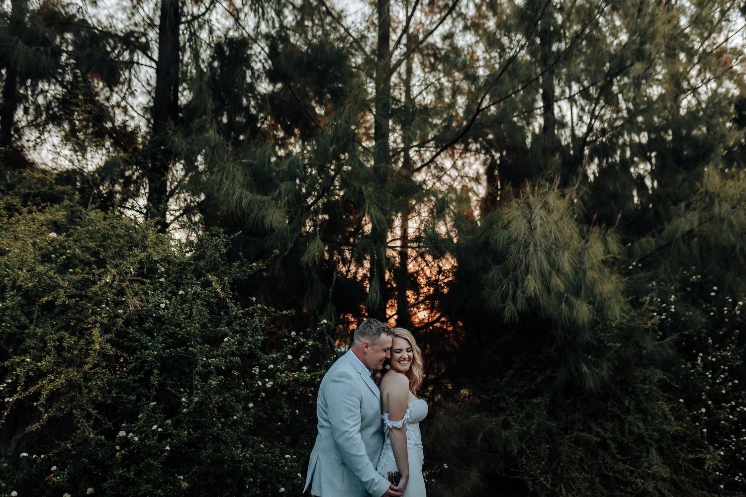 Newlywed couple shares intimate moment in forest setting during sunset.