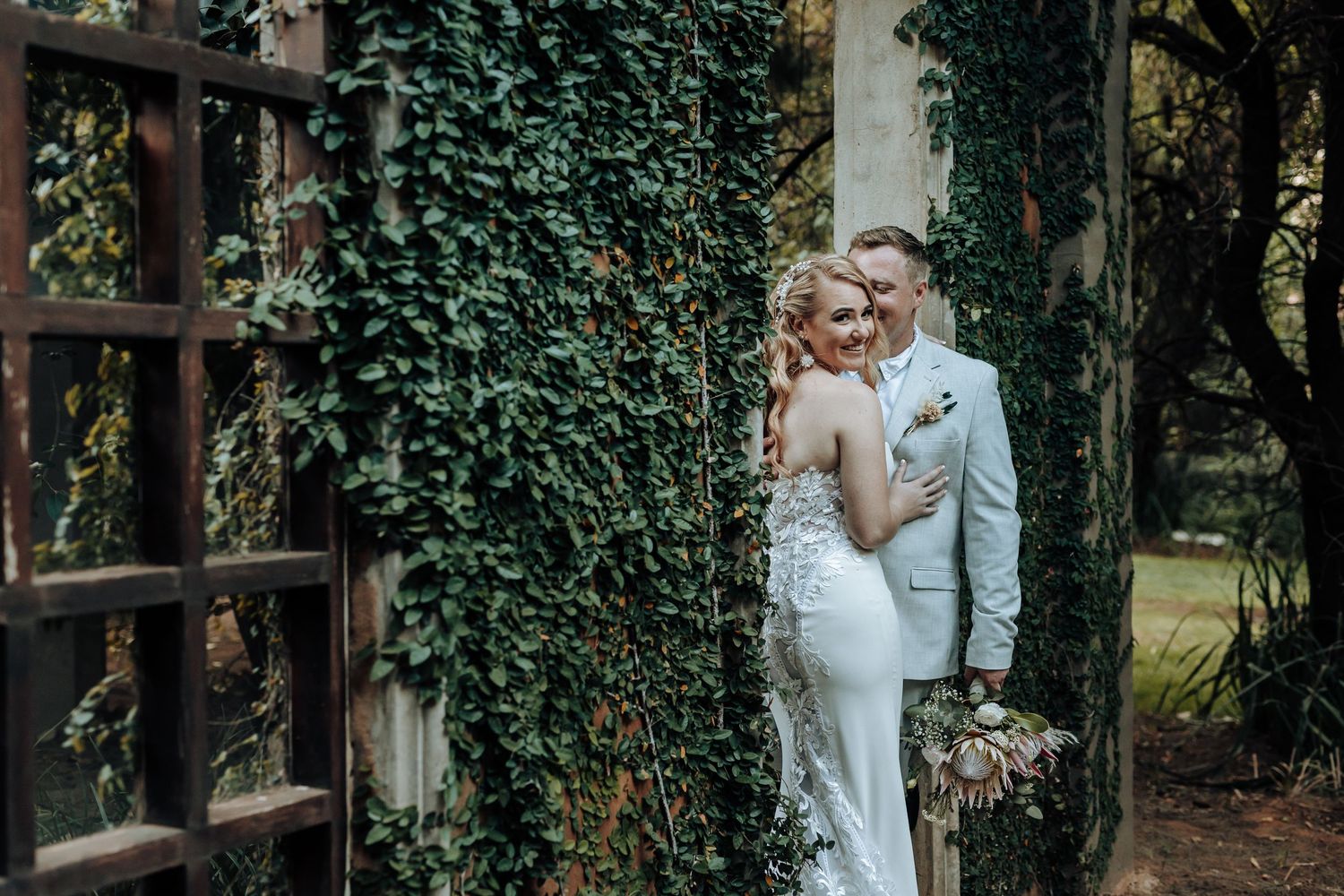 A romantic wedding couple poses against an ivy-covered wall in an elegant garden setting.