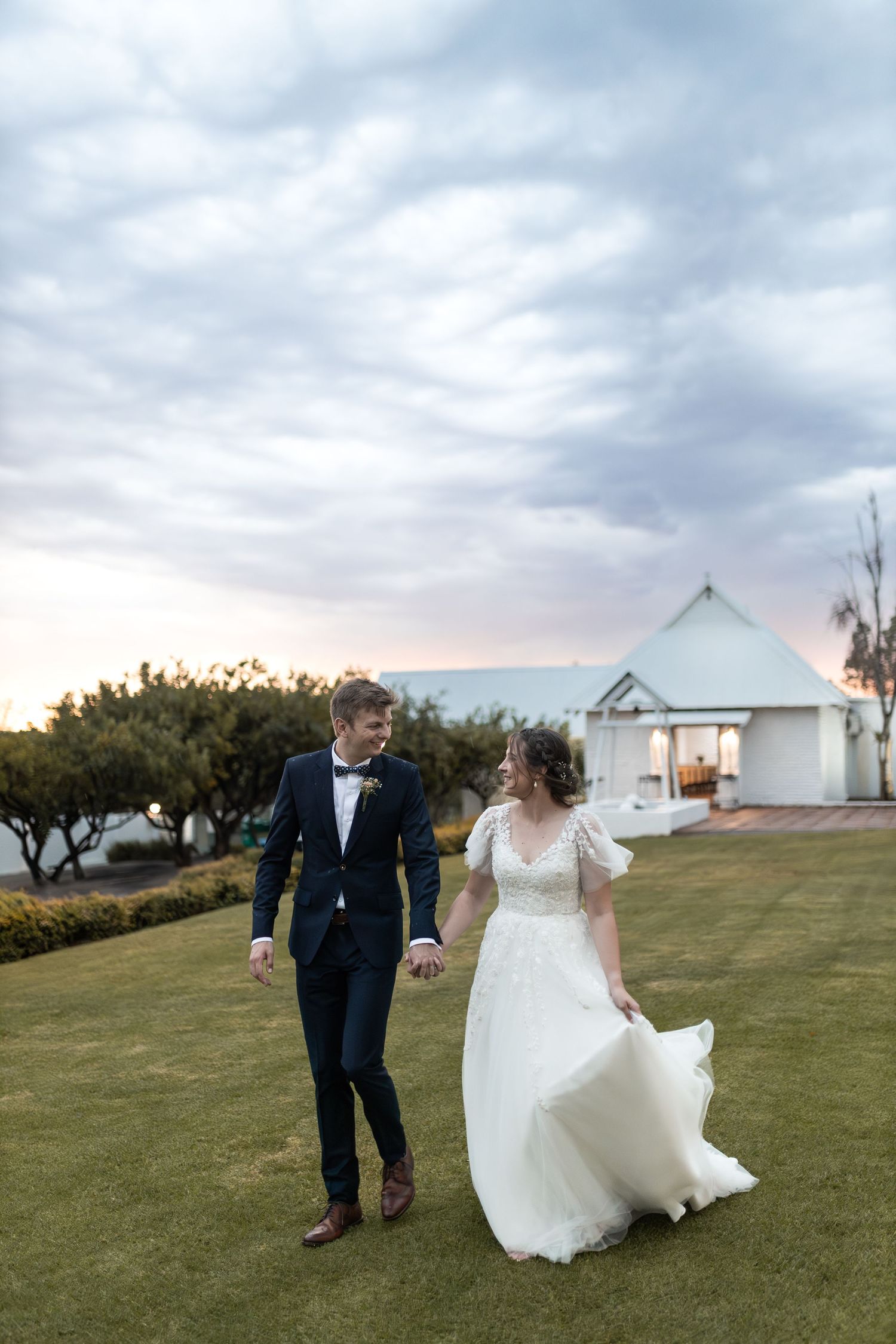 Wedding couple strolling on lawn with flowing dress and dramatic cloudy sunset sky.