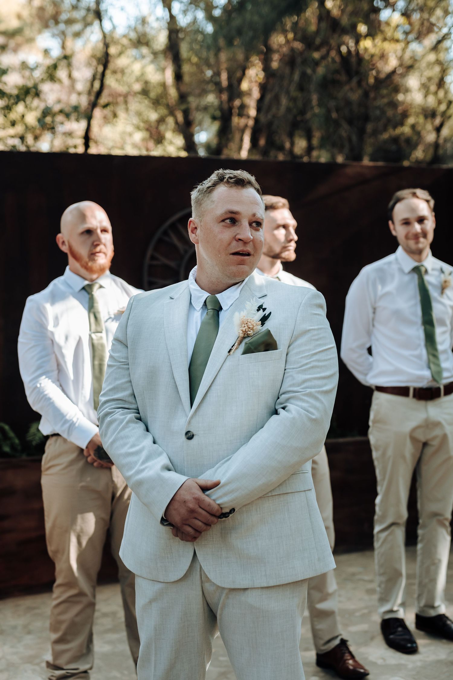 Groomsmen in light suits and green ties stand during an outdoor wedding ceremony.