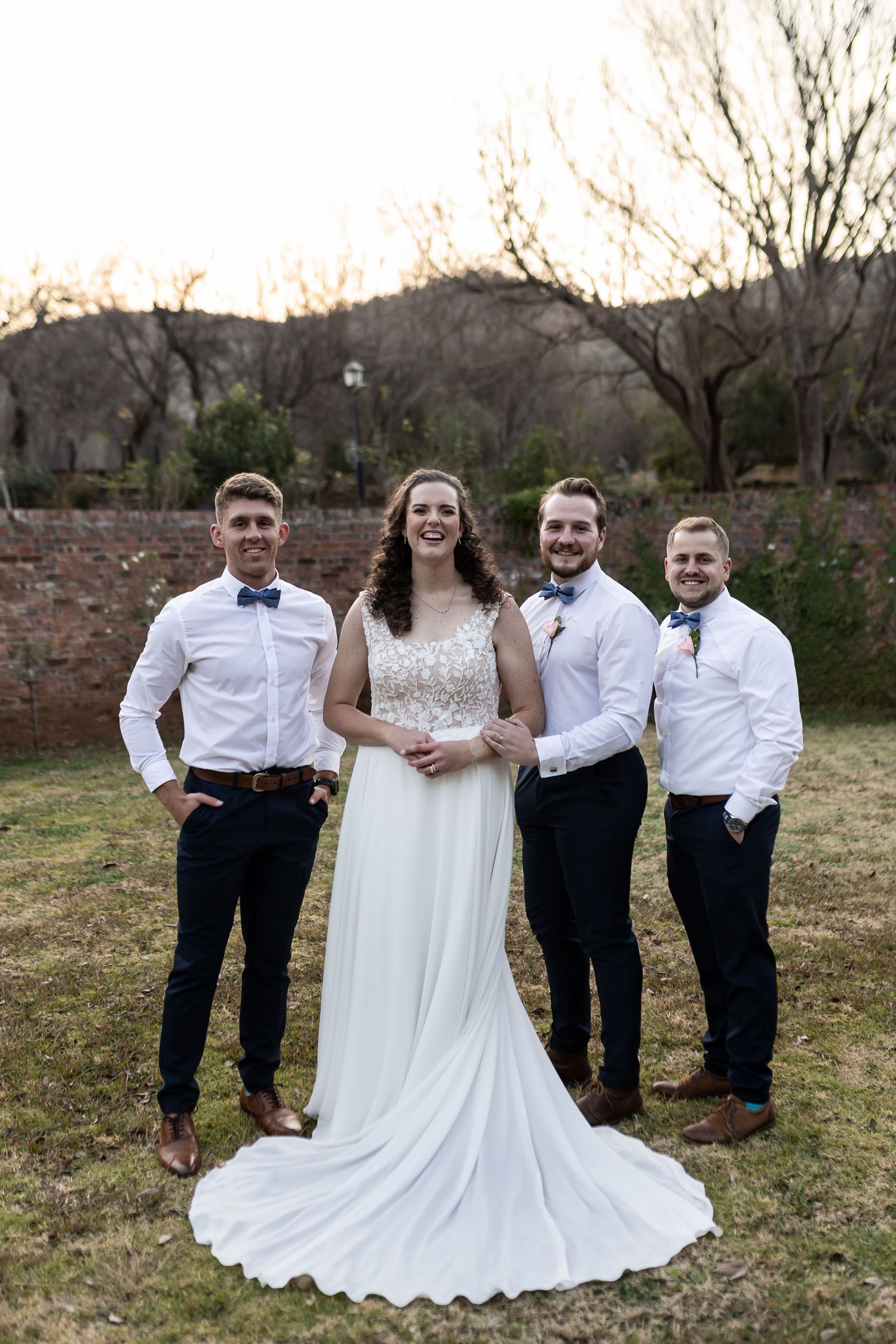 Wedding party in formal attire poses outdoors against brick wall and bare winter trees at sunset.
