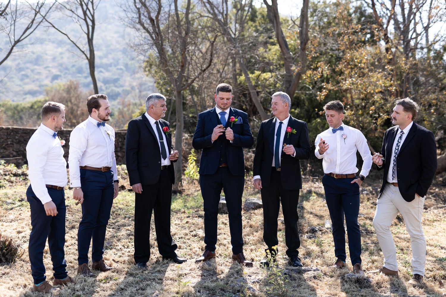 A group of seven men in formal attire stand together outdoors on a sunny autumn day.