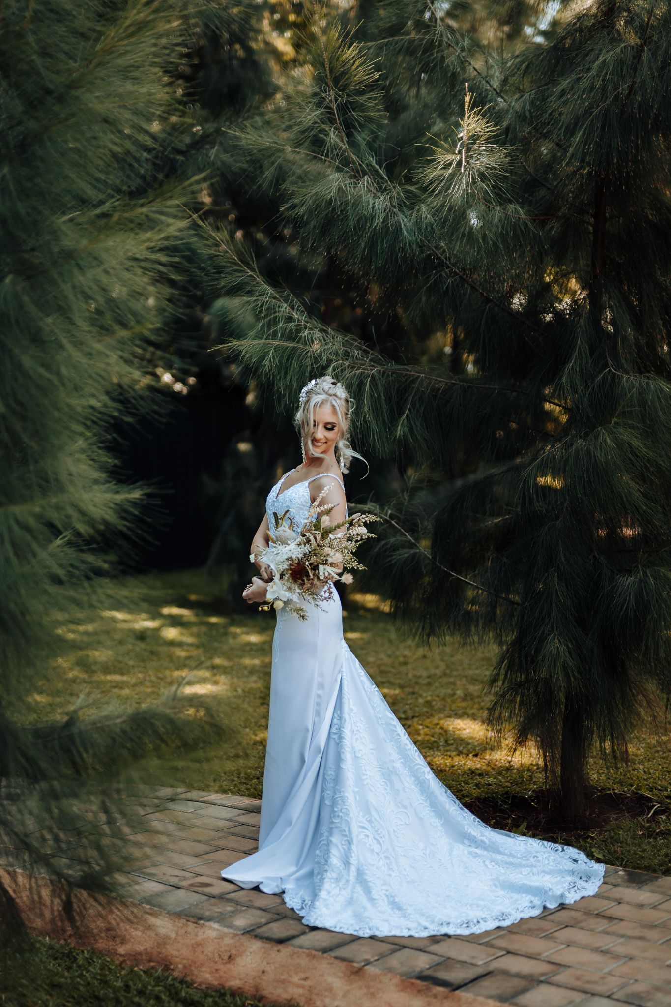 Elegant lace wedding gown with flowing train photographed in a garden setting with pine trees and dappled sunlight.