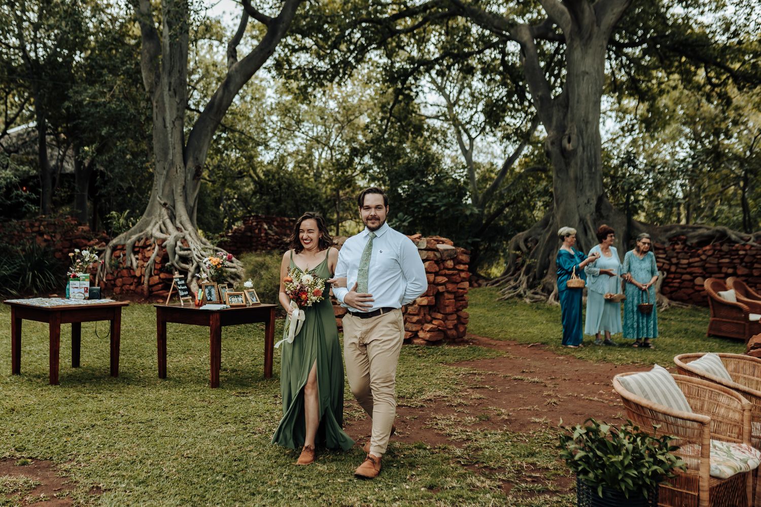 Wedding ceremony processional on outdoor garden path with rustic wooden furniture.