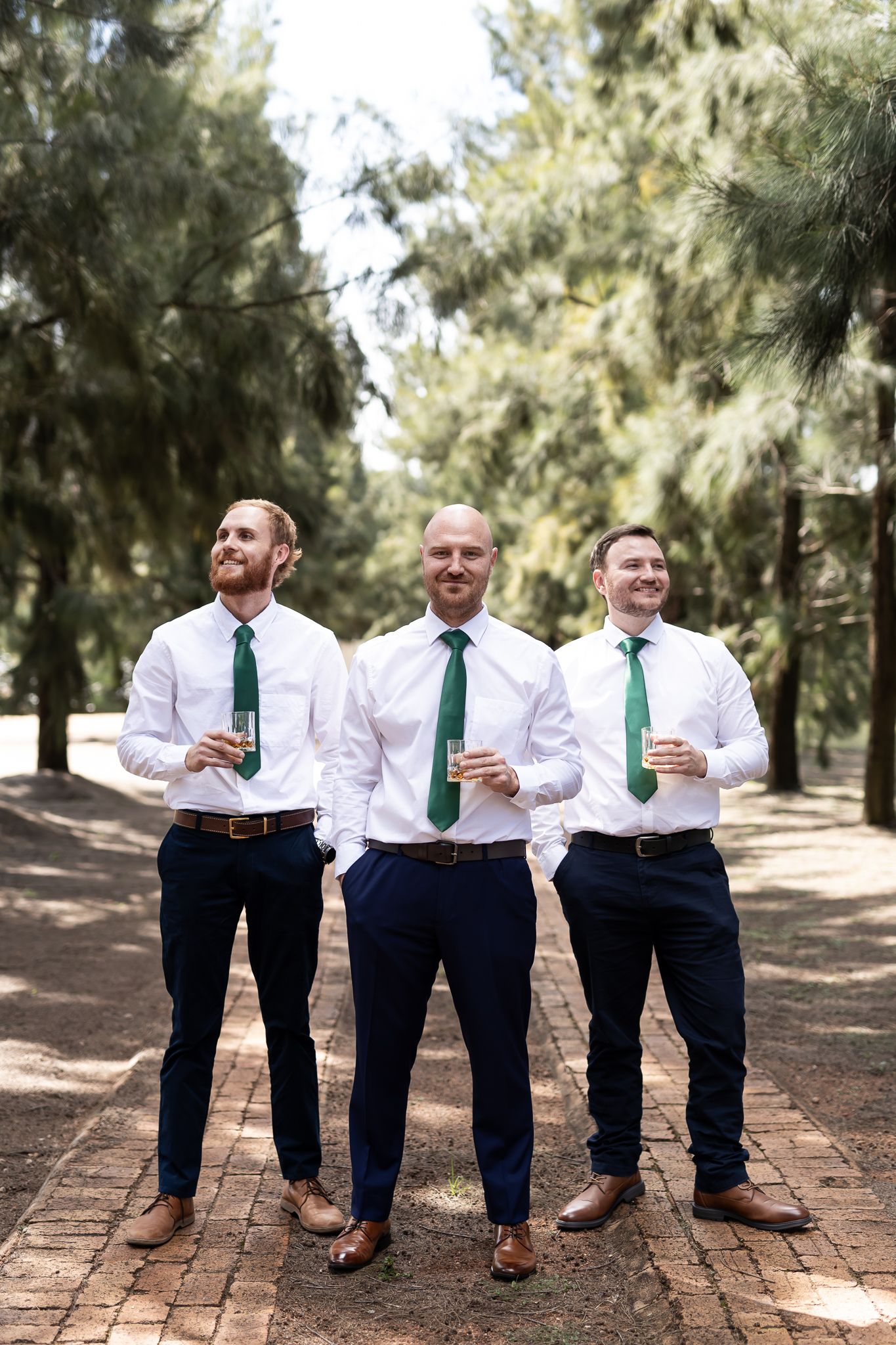 Three men in coordinated white shirts and green ties stand together on a brick pathway surrounded by pine trees.