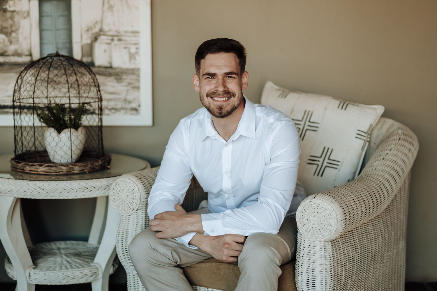 Portrait of person in white shirt sitting on cream armchair next to decorative birdcage.