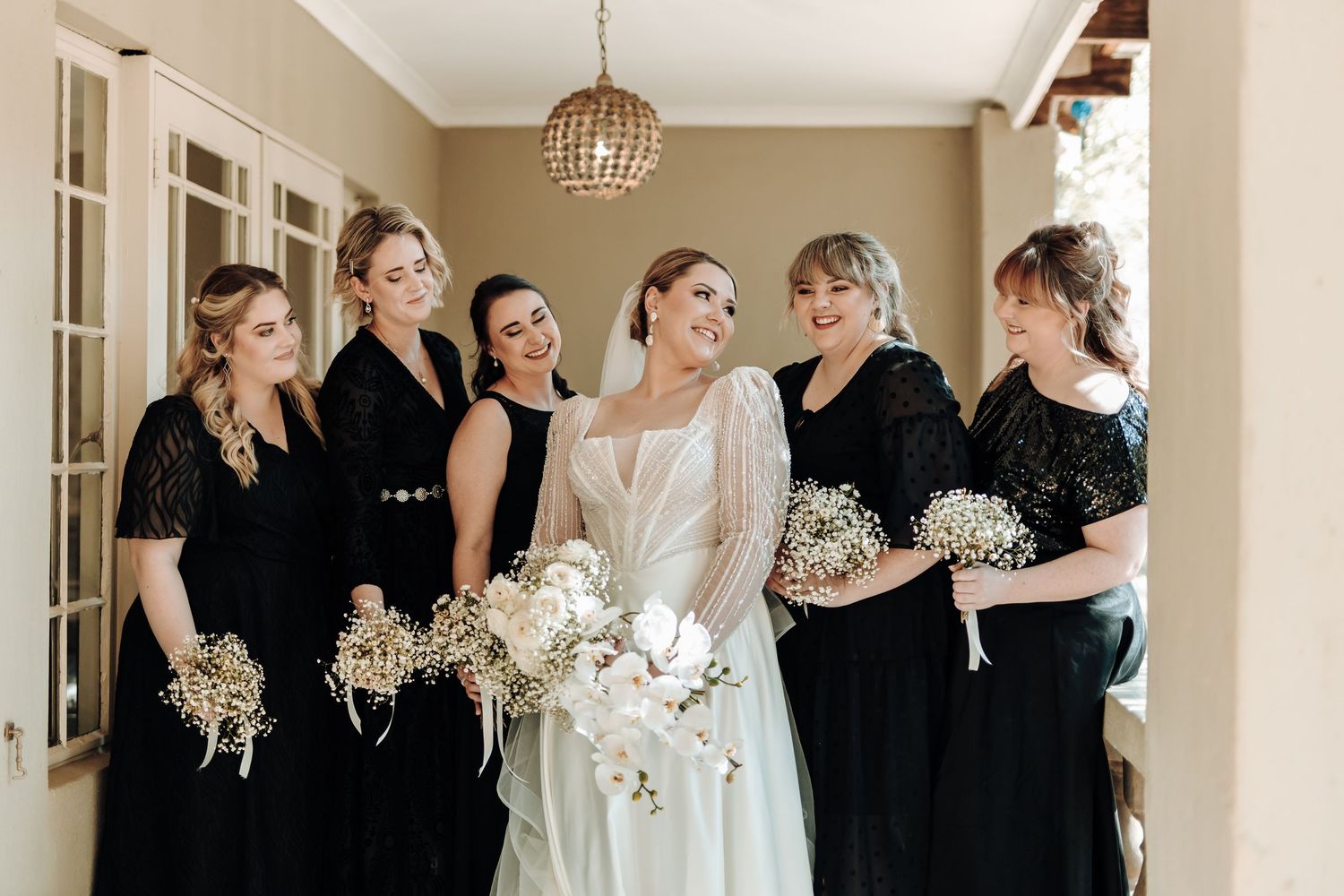 Bridal party in black dresses pose with bride in white gown holding matching bouquets.