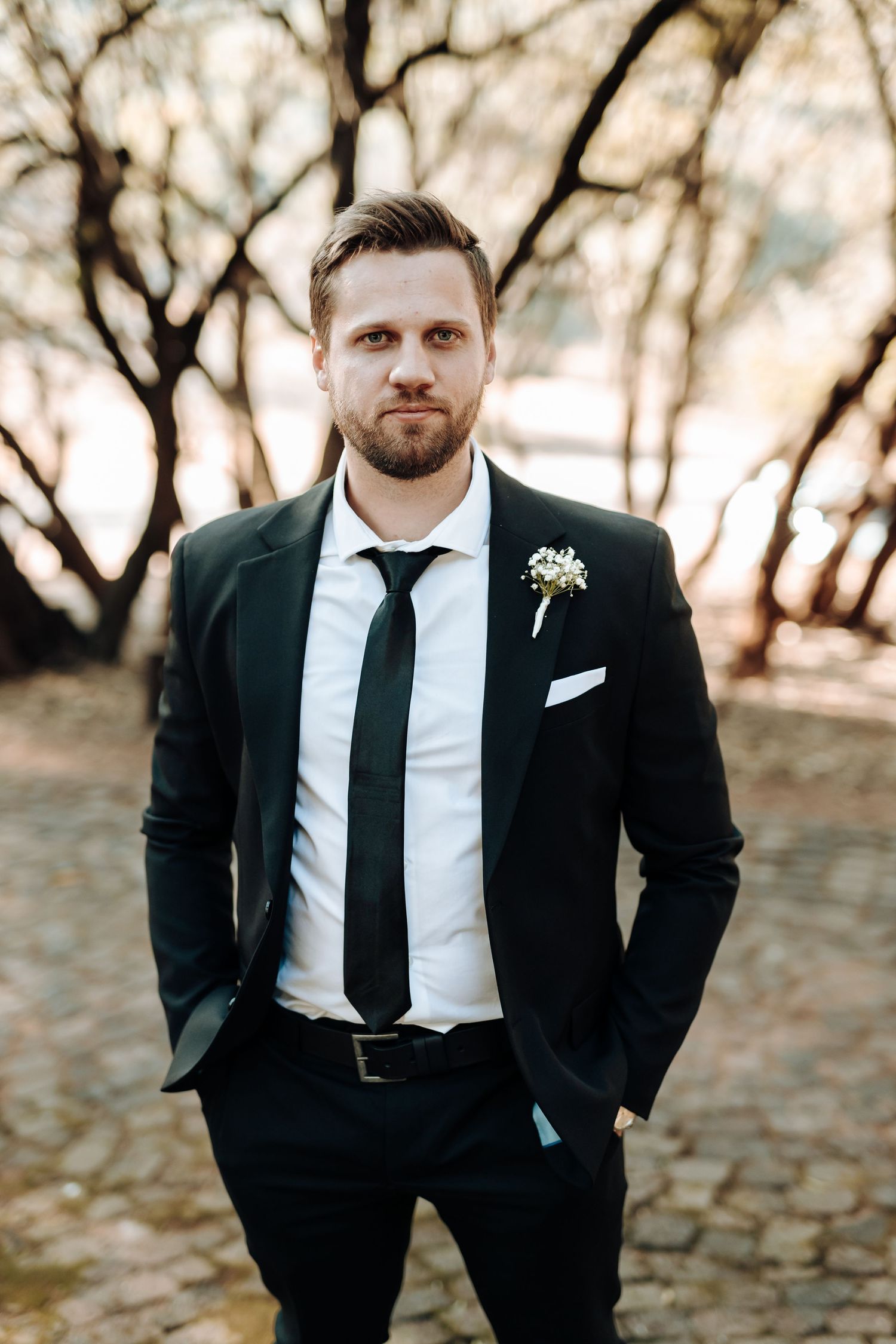Groomsman in black suit and tie poses against autumn trees with boutonniere.