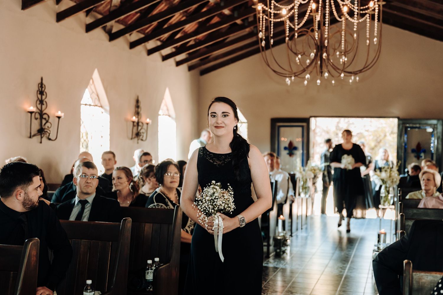 Bridesmaid in black dress walks down church aisle holding small bouquet.