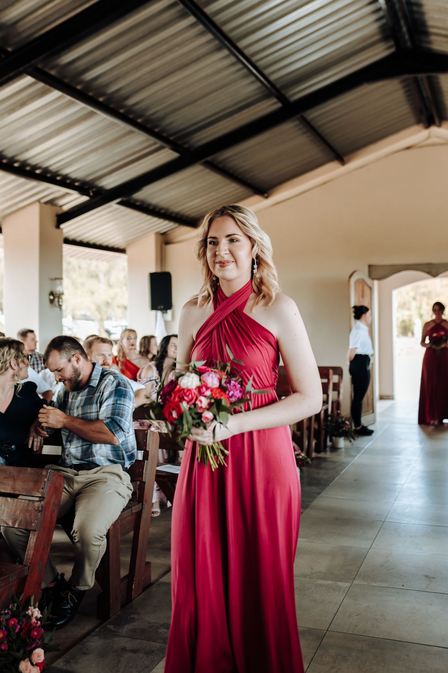 Bridesmaid in halter-style red gown holds pink bouquet during ceremony processional.