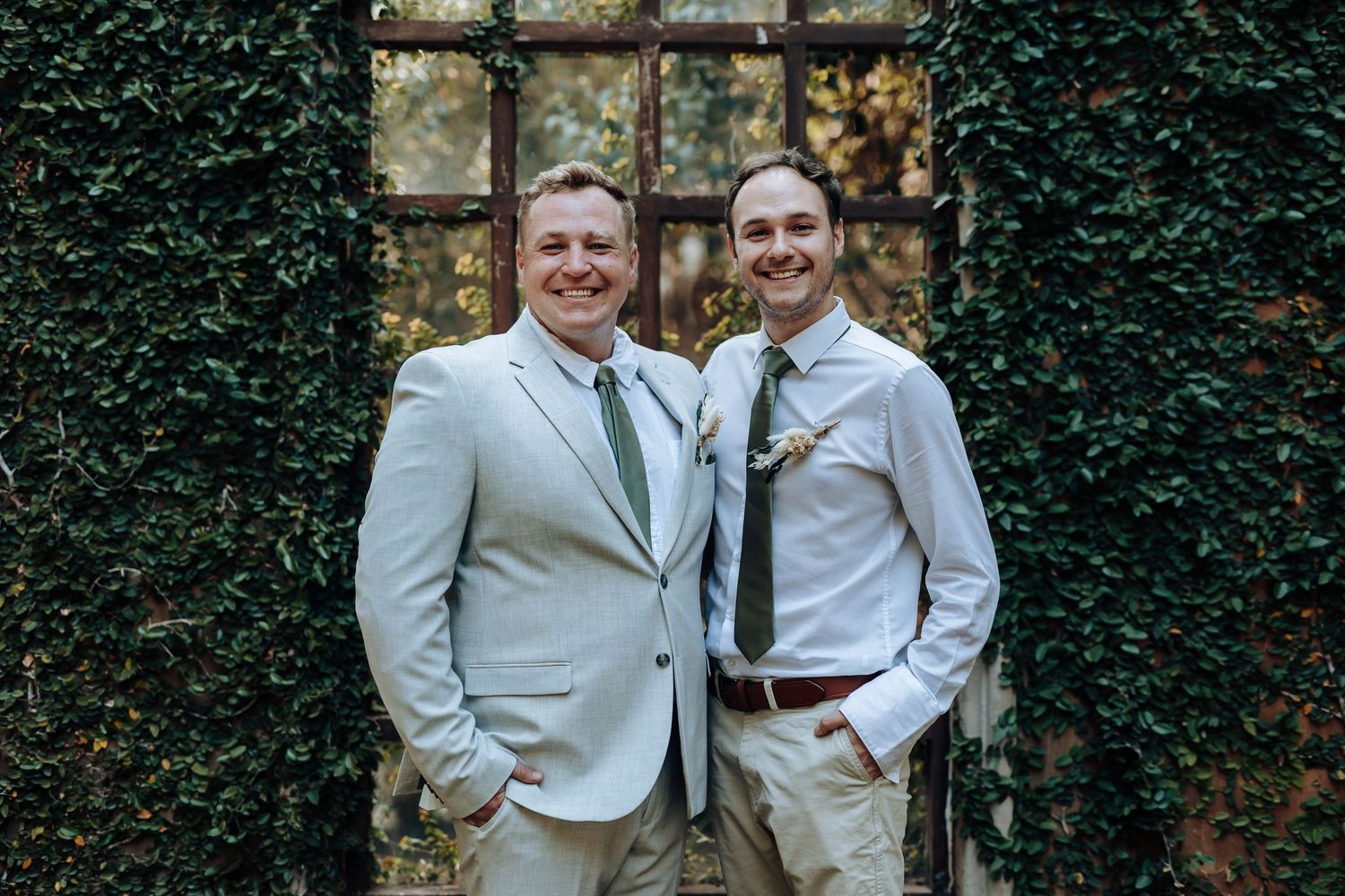 Two people in light colored suits pose together in front of an ivy-covered wall and window frame.