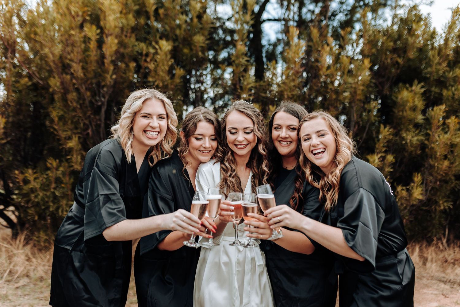 Wedding party wearing black robes toasts with champagne flutes in a joyful outdoor celebration.