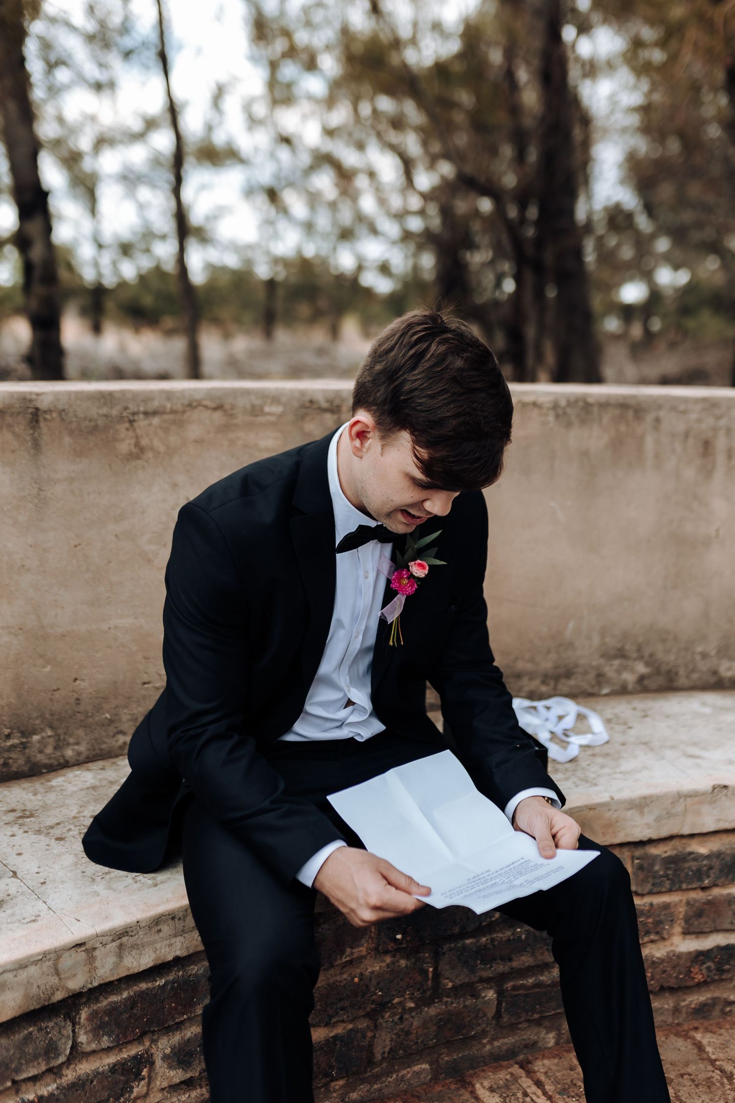 A person in formal attire reads papers while seated on a concrete bench outdoors.