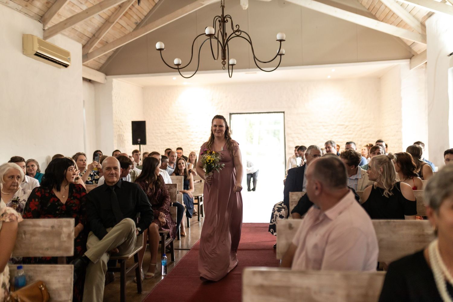 Wedding attendant in sage gown walks processional aisle in chapel filled with guests.