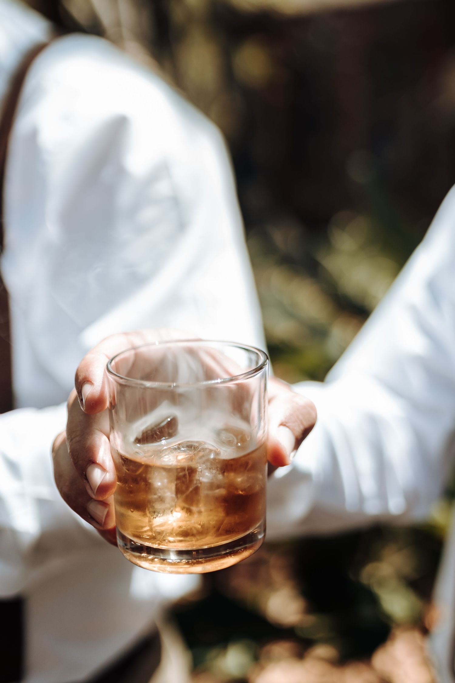 Hand holding a crystal glass of whiskey with ice against white shirt sleeve.