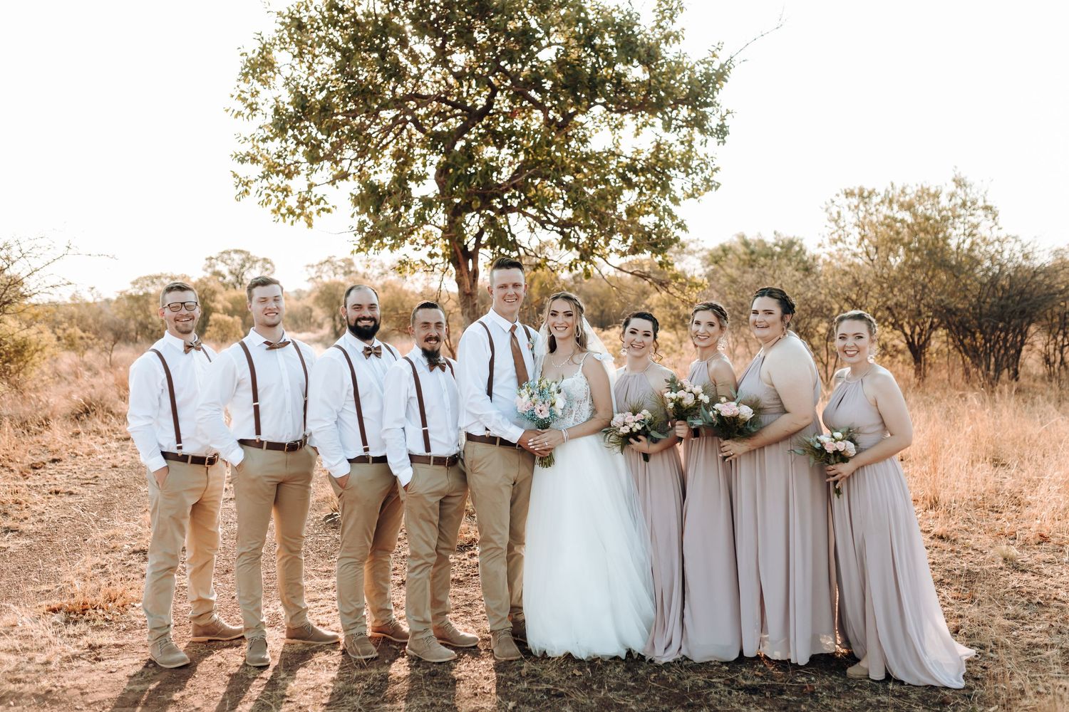 Wedding party in khaki and blush attire posing together under a tree in rustic outdoor setting.