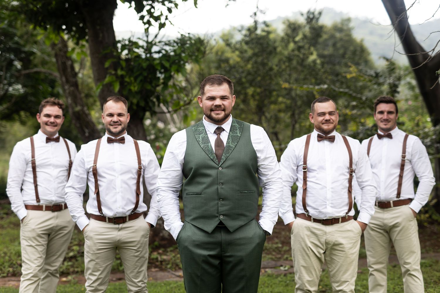 Wedding party in coordinated outfits with suspenders poses in an outdoor garden.