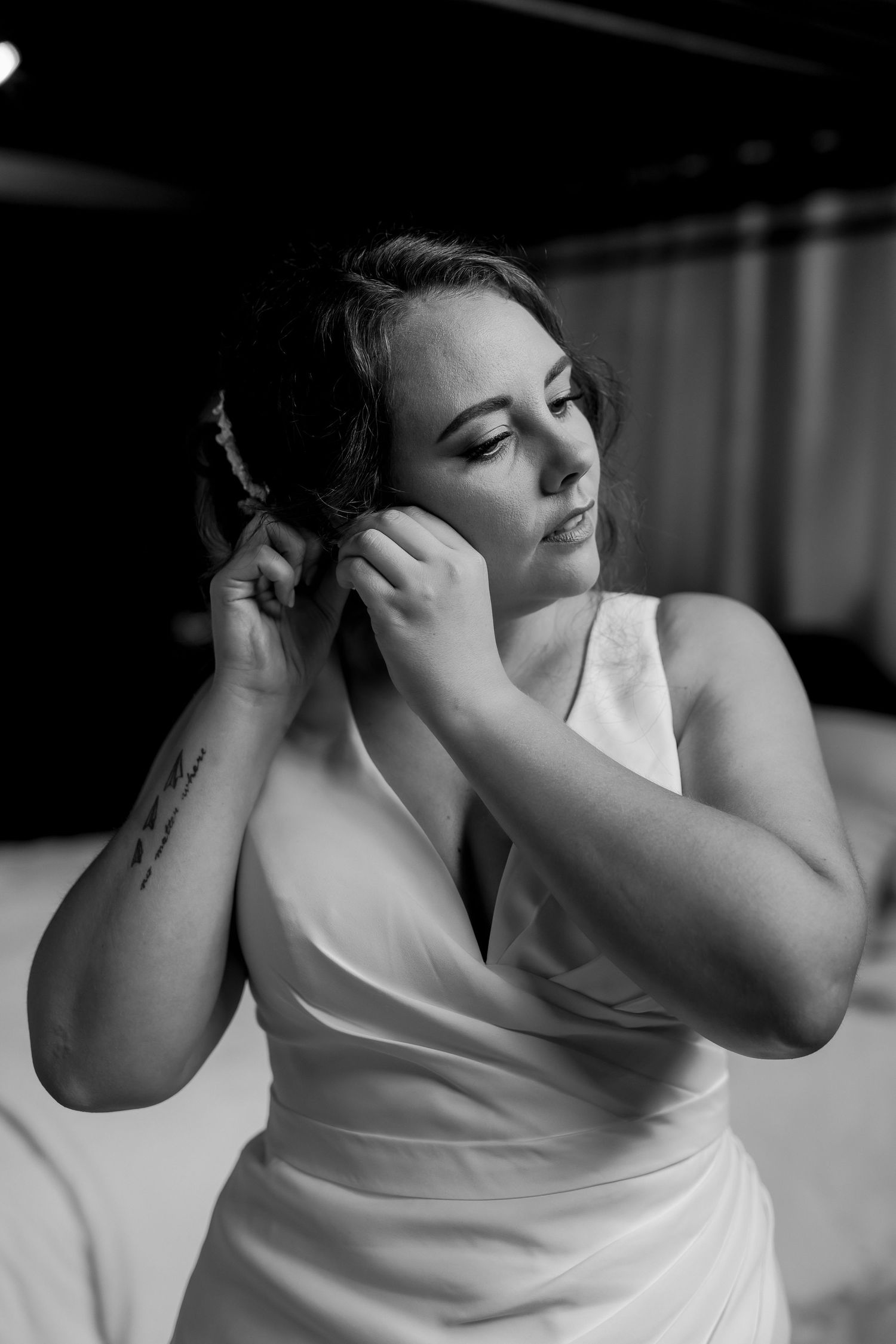 Bride in white satin dress adjusts hair in dramatic black and white portrait.