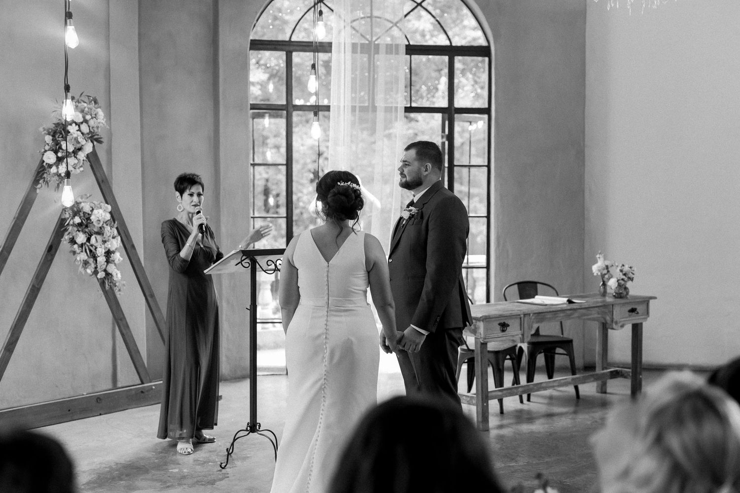 Indoor wedding ceremony in elegant venue with arched windows and floral decorations.