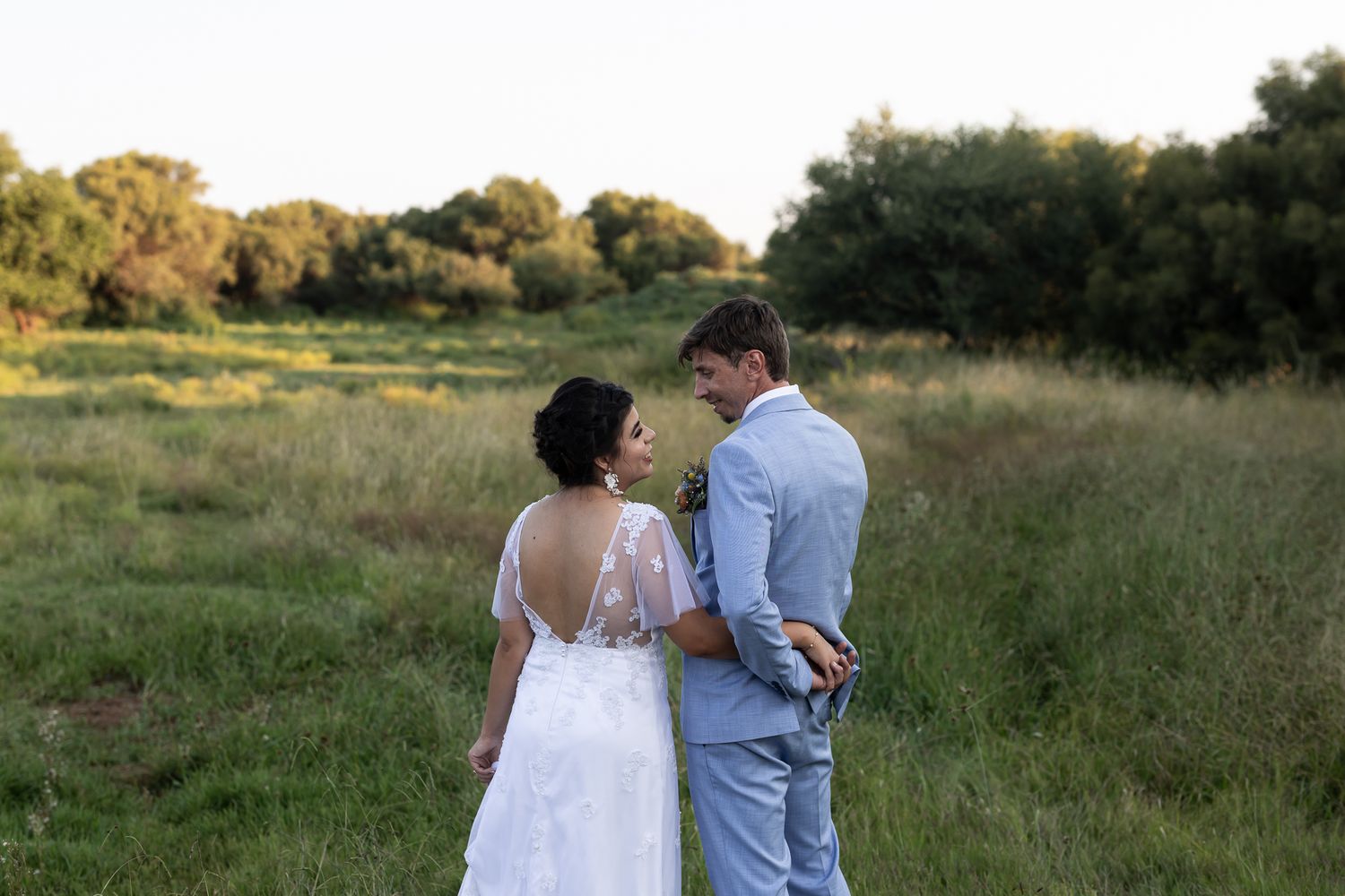 A couple walks hand in hand through a meadow with trees in the background.
