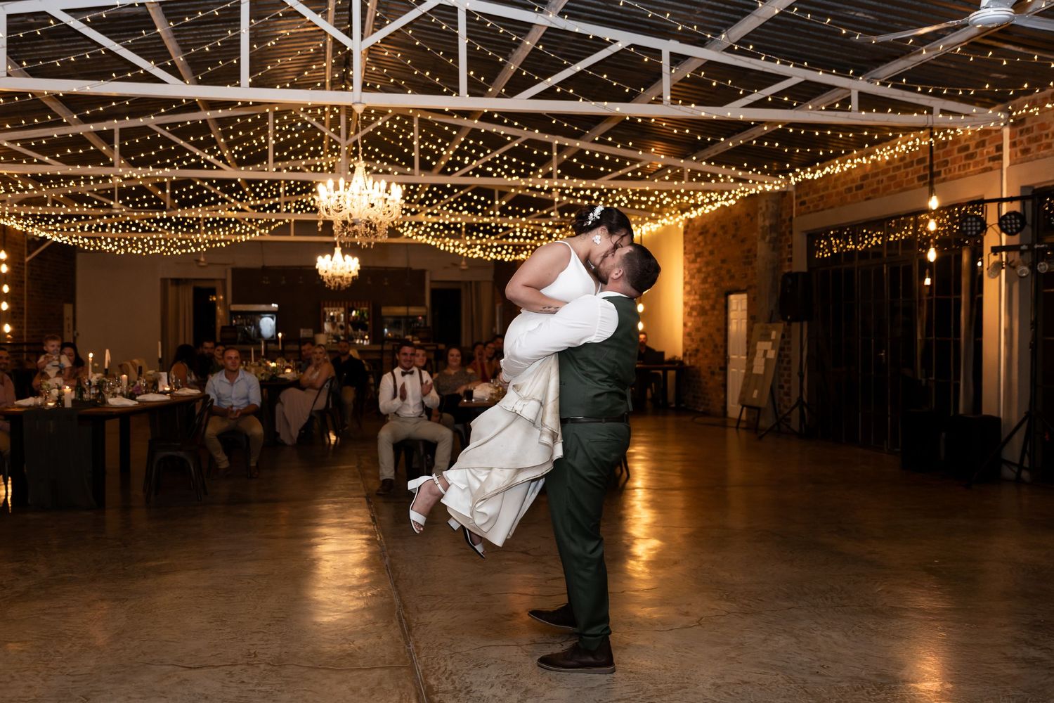 A playful dance lift moment in an elegant reception hall with string lights overhead.