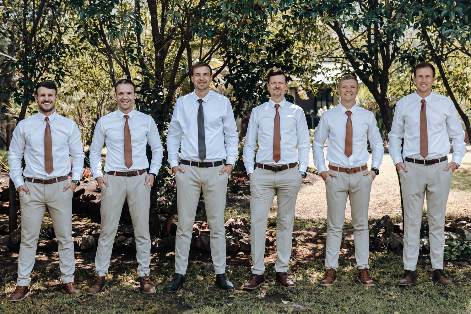 Wedding party in light gray suits and brown ties standing in line against trees.