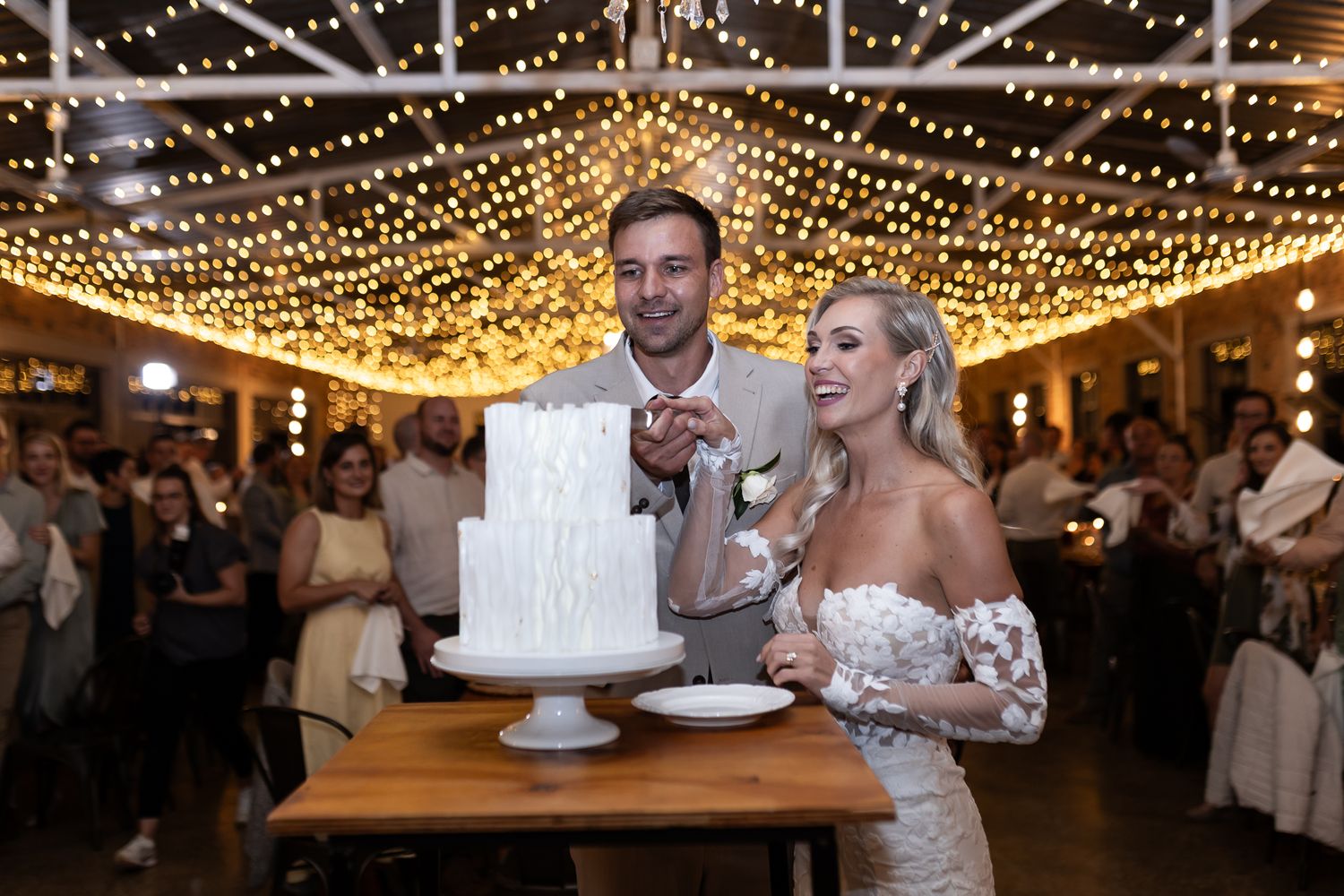 Wedding couple cutting their elegant white tiered cake under warm string lights.