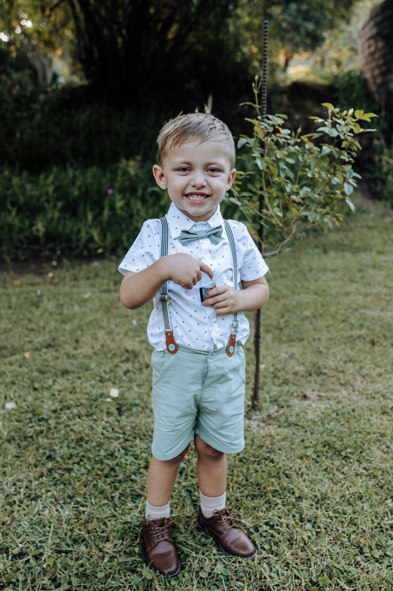 Smiling child in formal wear with mint green suspenders stands outdoors.