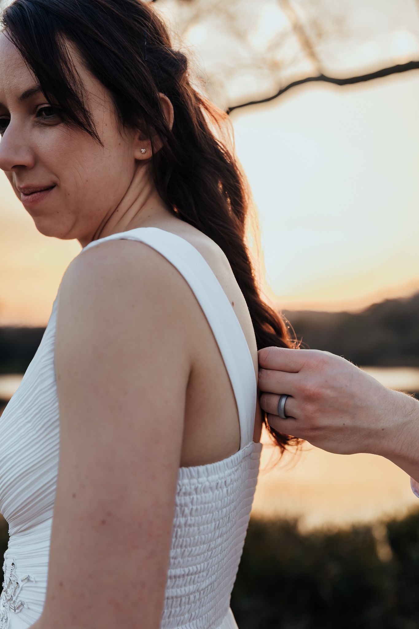 A cropped view of someone adjusting the zipper on a white one-shoulder wedding dress at sunset.